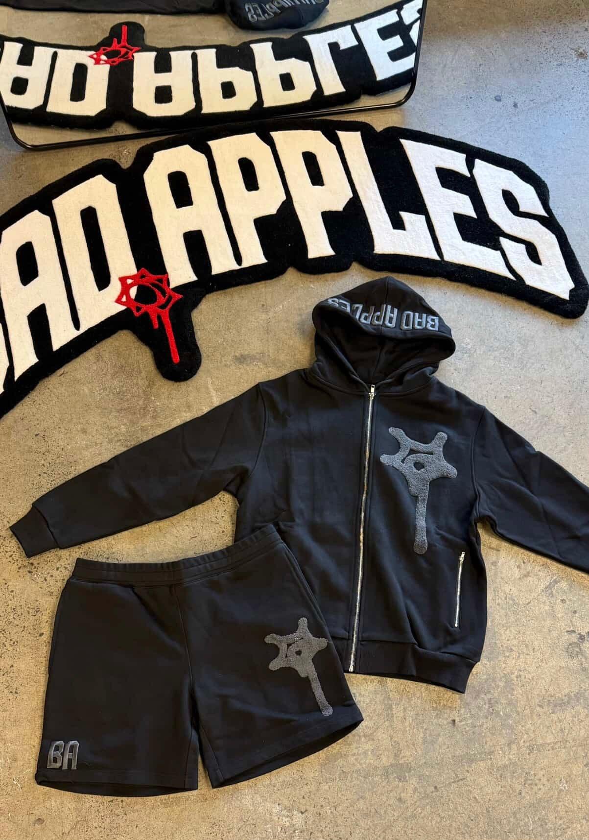 Black handmade rug with 'BAD APPLES' text and matching black hoodie and shorts set on floor