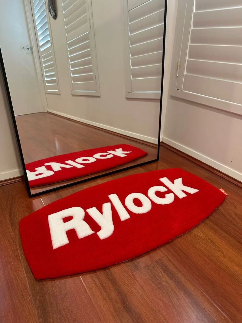 Red handmade tufted rug with white 'Rylock' text on wooden floor beside a mirror
