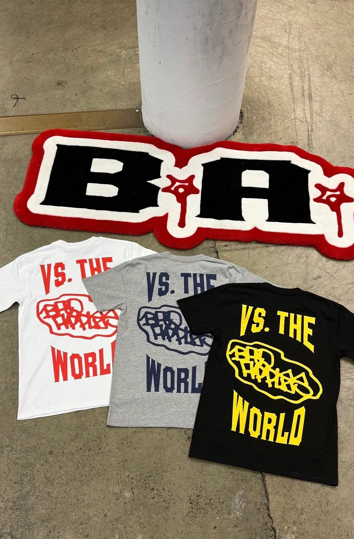 Handmade logo rug with bold black, white, and red design beside three 'VS. THE WORLD' graphic t-shirts in white, grey, and black on concrete floor.