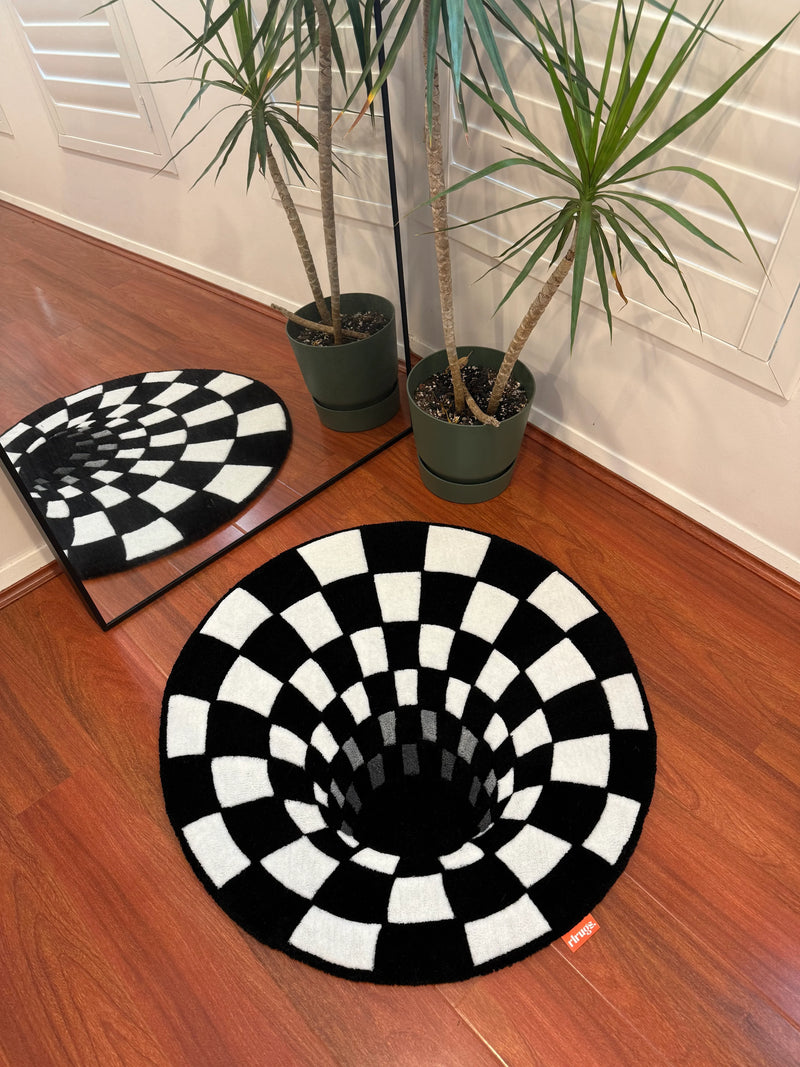 Handmade black and white checkered optical illusion rug on wooden floor near potted plant