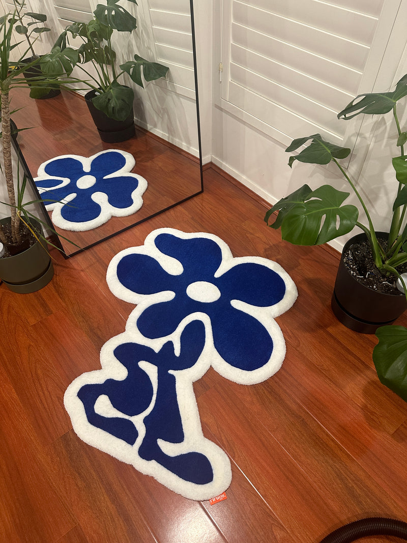 Handmade blue flower tufted rug with white trim on wooden floor, near mirror and plants.