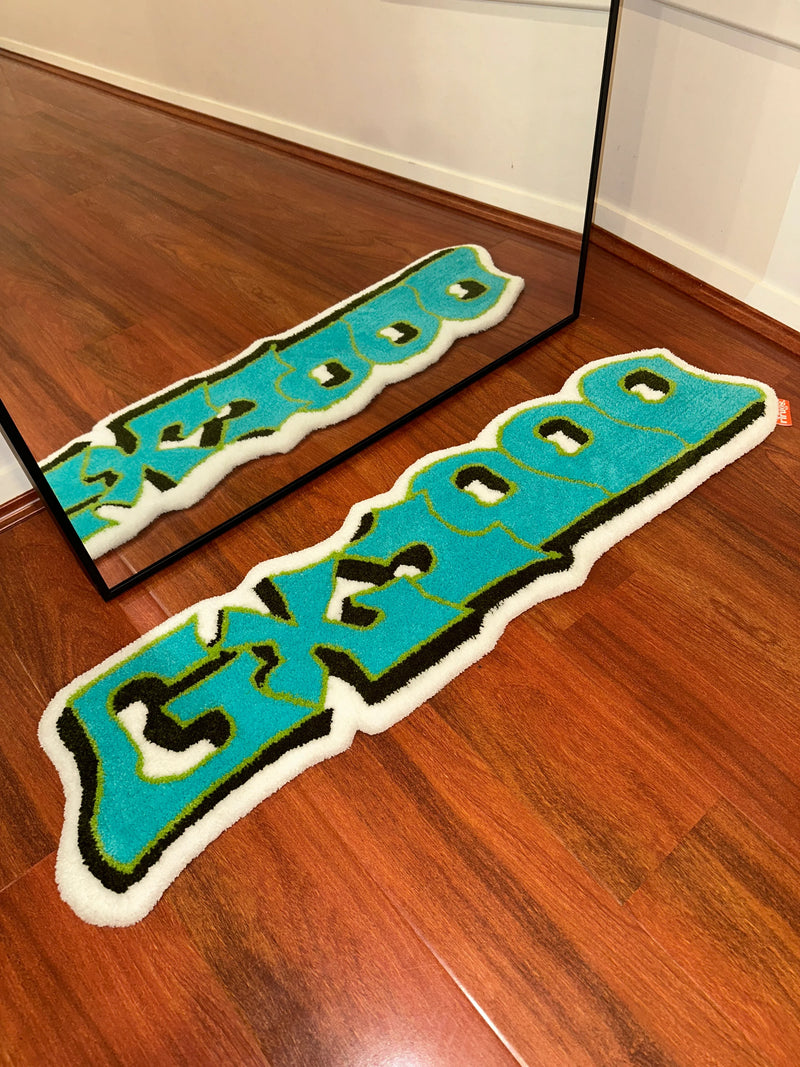 Handmade tufted rug with blue graffiti-style text on wood floor, reflected in a mirror.