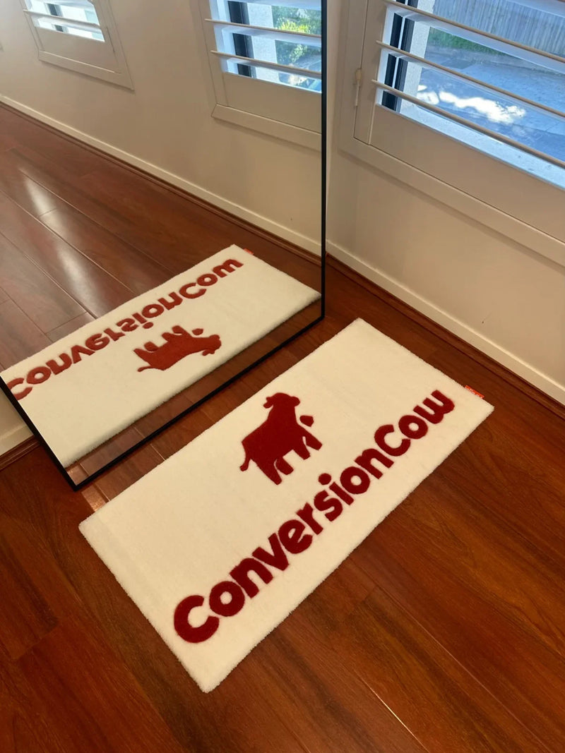 White custom rug with red text 'ConversionCow' and cow graphic on wooden floor by window and mirror